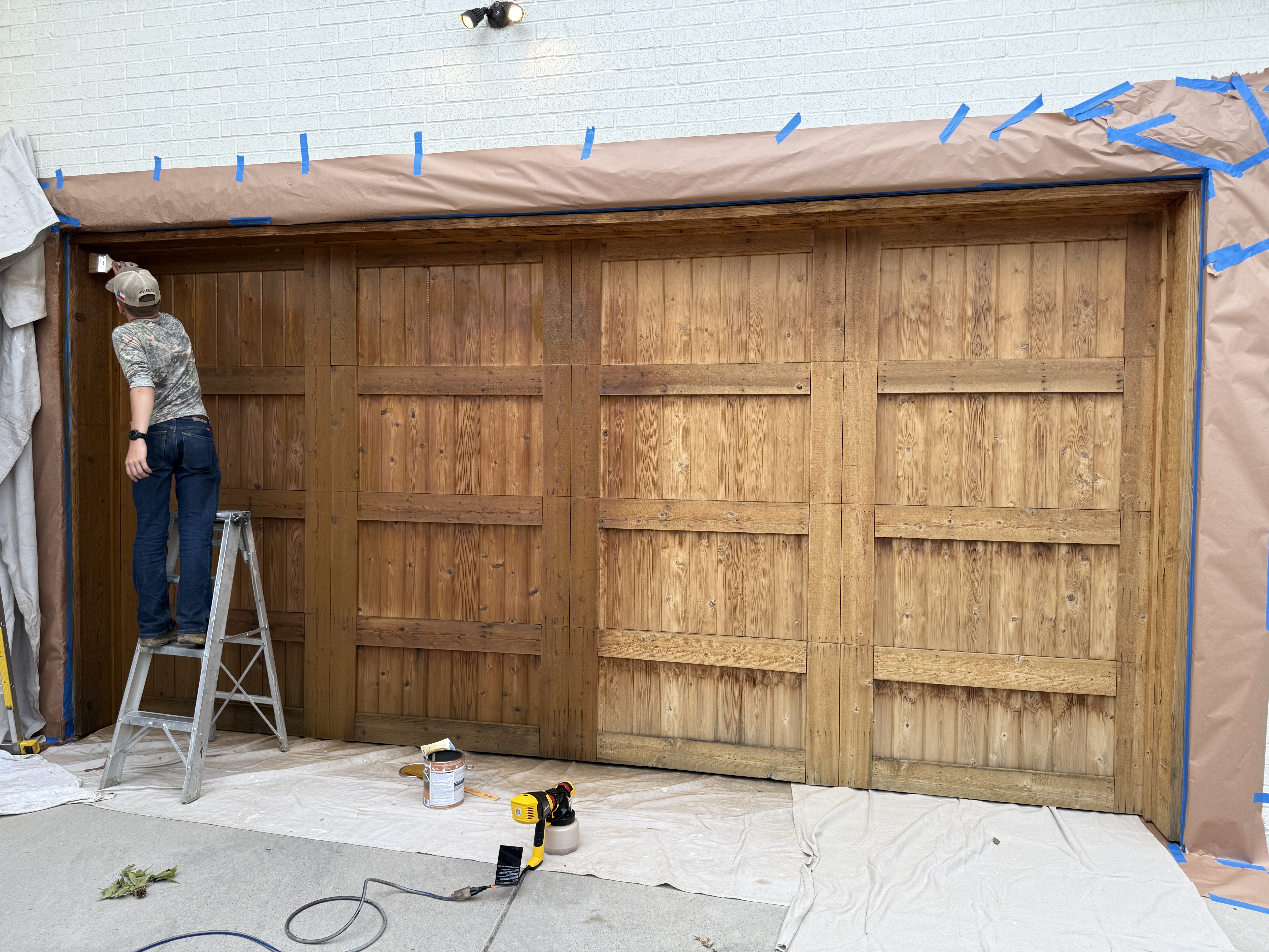 A weathered wood garage door being stained, the faded gray still showing near the top