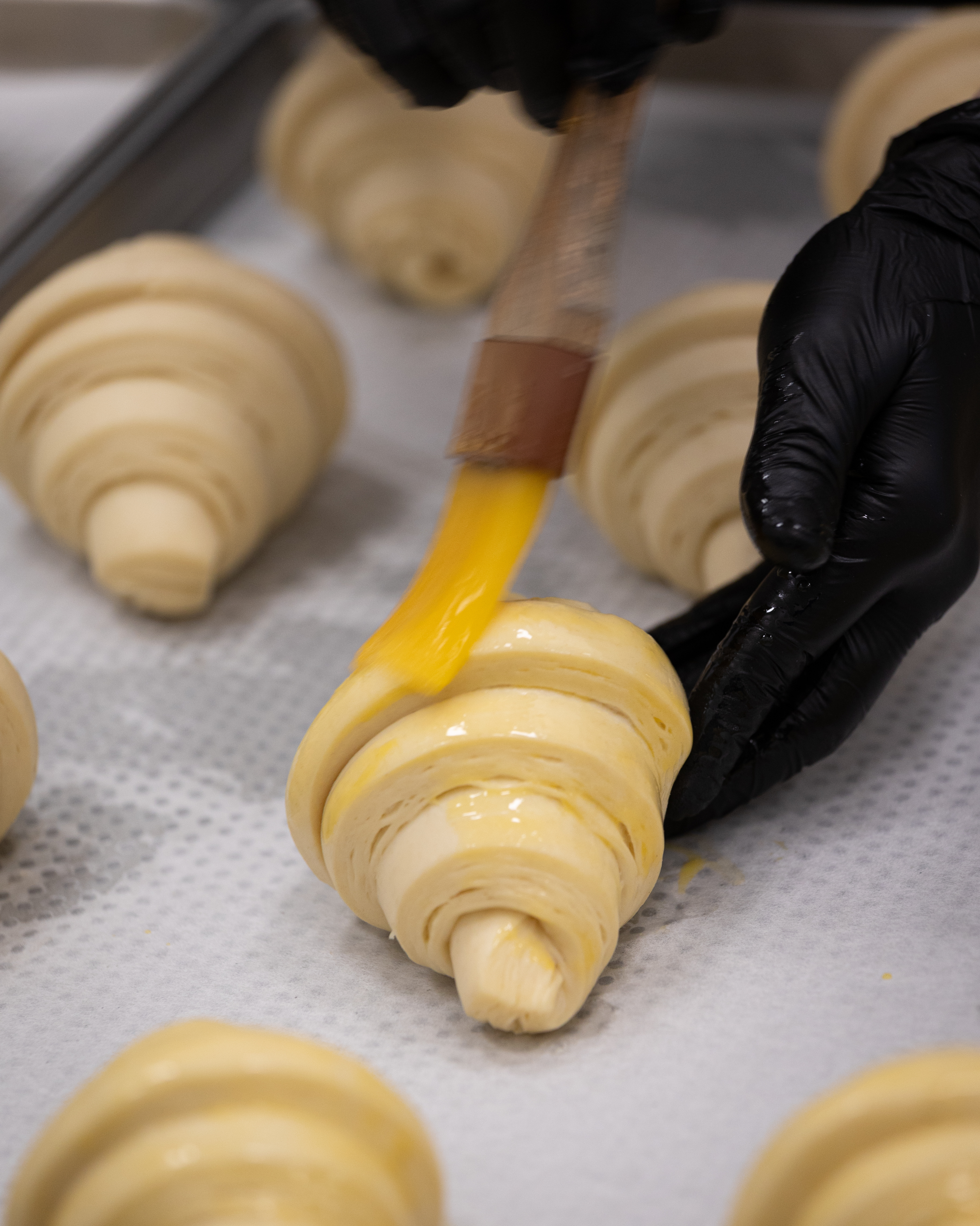 A baker brushing egg wash onto rows of raw croissants